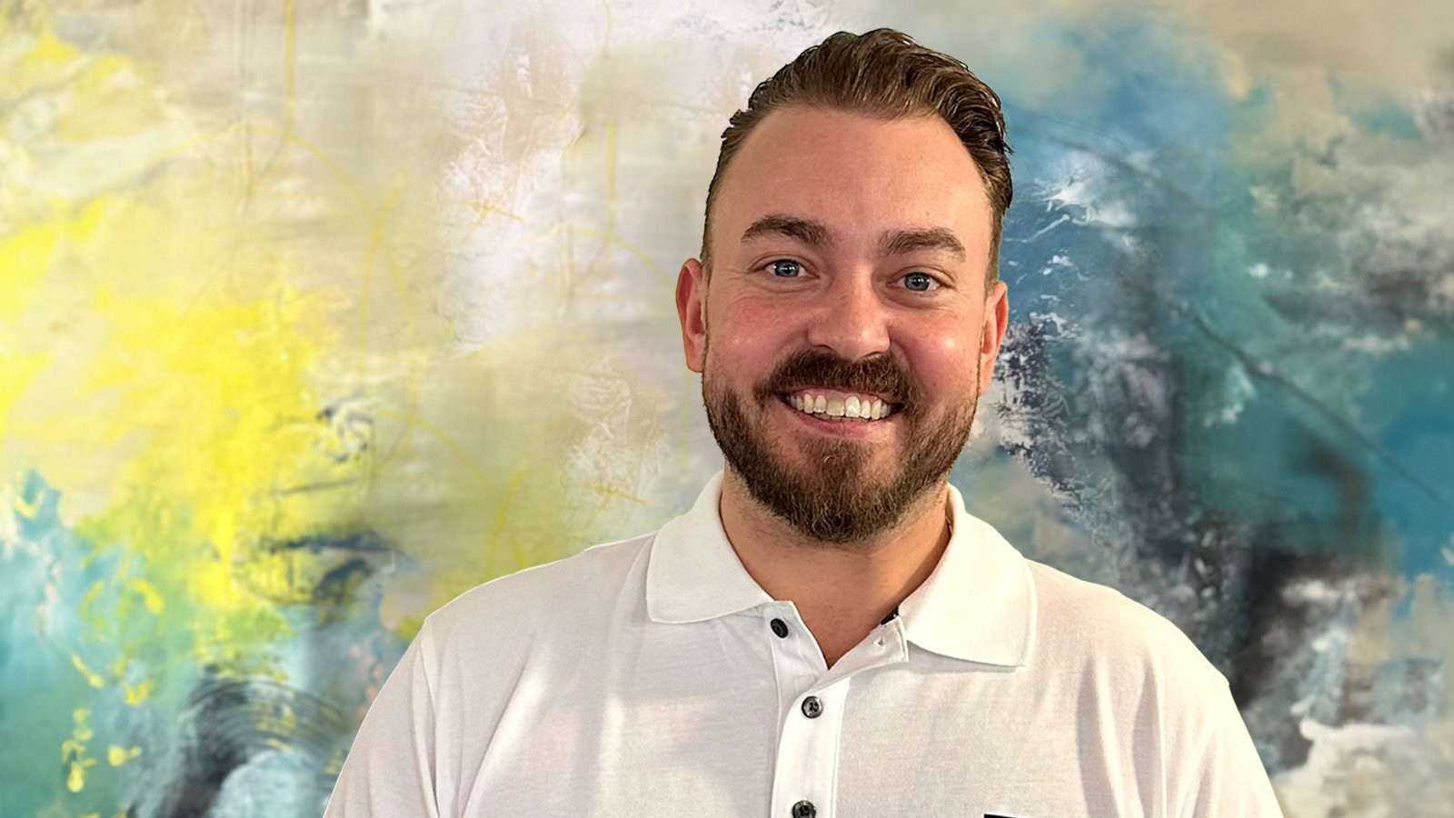 Lächelnder Mann in einem weißen Hemd vor einem abstrakten Gemälde mit gelben und blauen Farbklecksen.